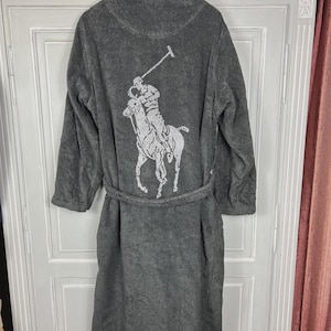 Op de afbeelding: Een grijze badjas van badstof met een sjaalkraag en een bijpassende riem. De achterkant van de badjas heeft een wit polospelerlogo. De badjas hangt aan een witte deur.