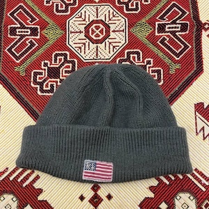 May include: A dark gray knit beanie with a folded brim. A small patch featuring the American flag is sewn on the front. The hat is displayed on a patterned fabric with red, white, and gold geometric designs.