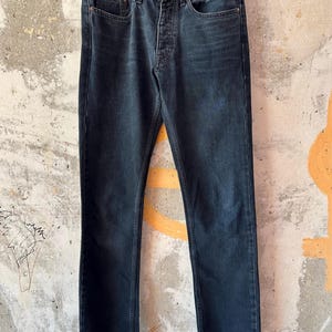 May include: Dark wash denim jeans with a button fly and classic five-pocket design. The jeans are cuffed at the ankles and hang from a white hanger. The brand name "ACNE STUDIOS" is visible on the waistband.