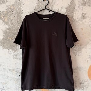 May include: A black crew neck t-shirt hanging on a black hanger. The shirt has short sleeves and a small embroidered "folk" logo on the chest. The background is a textured, off-white wall.