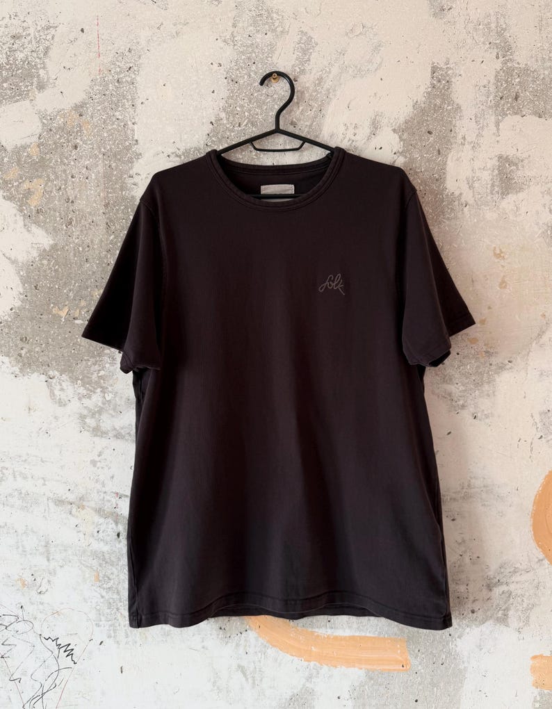May include: A black crew neck t-shirt hanging on a black hanger. The shirt has short sleeves and a small embroidered "folk" logo on the chest. The background is a textured, off-white wall.