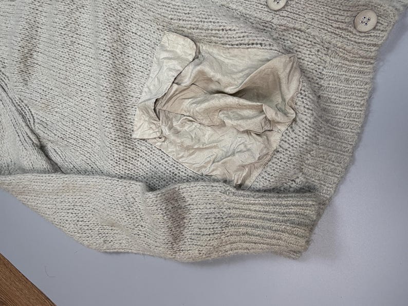 Op de afbeelding: Een lichtbeige gebreide trui met een grove, getextureerde weving en een grote, opgestikte zak van gladde contraststof aan de voorzijde. Het kledingstuk is voorzien van een geribbelde boord en twee ronde, lichtgekleurde knopen aan de bovenrand.