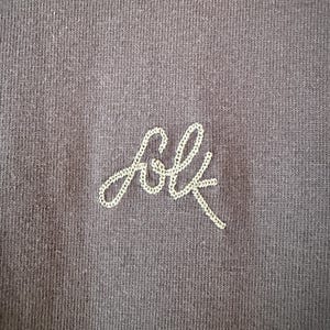 May include: Close-up of a dark gray fabric with the embroidered word "folk" in a cursive font. The embroidery is done in a light cream-colored thread, creating a subtle contrast against the darker background.