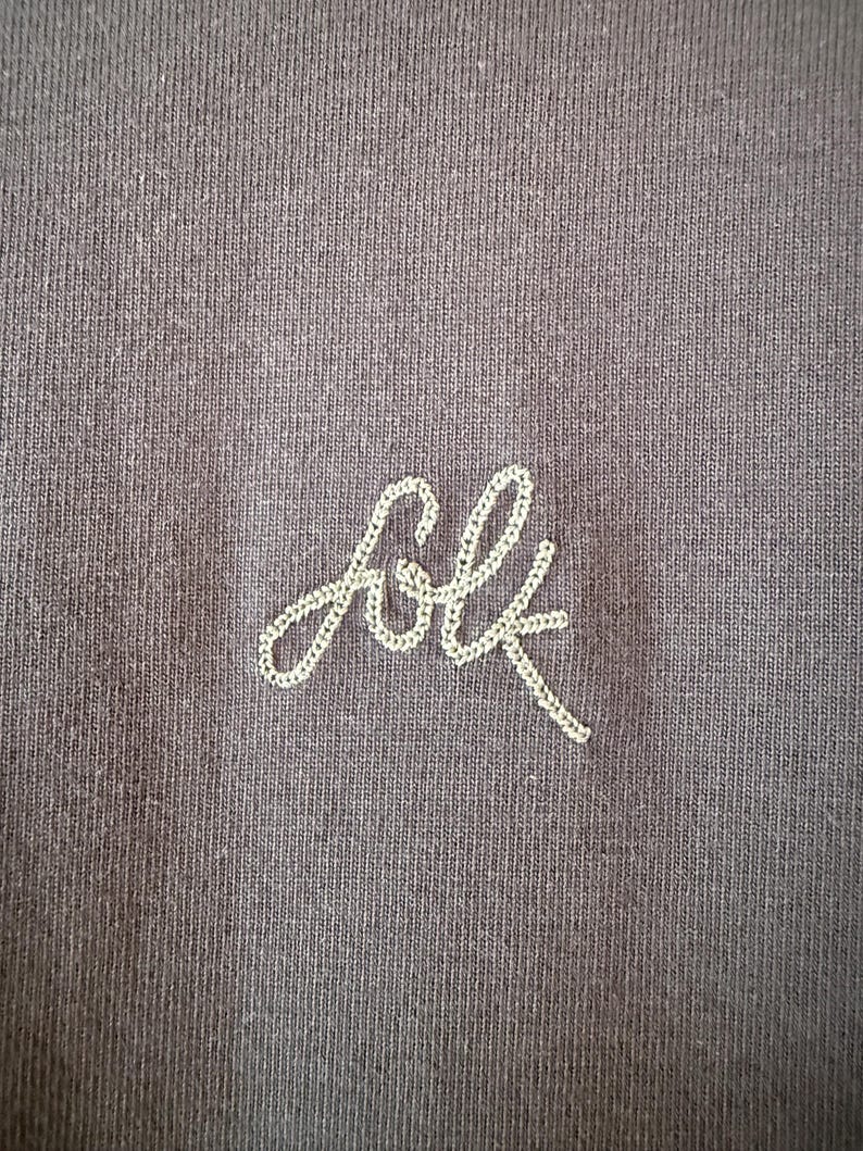 May include: Close-up of a dark gray fabric with the embroidered word "folk" in a cursive font. The embroidery is done in a light cream-colored thread, creating a subtle contrast against the darker background.