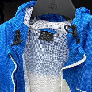 K&ouml;nnte beinhalten: Eine blau-wei&szlig; gemusterte Jacke mit wei&szlig;em Mesh-Futter. Die Jacke hat einen Rei&szlig;verschluss und eine Kapuze. Das Etikett auf der Jacke lautet "Oakley XL/TG".