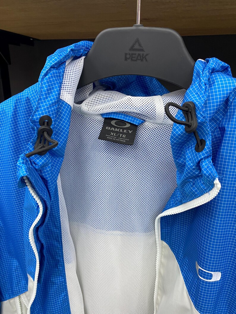 K&ouml;nnte beinhalten: Eine blau-wei&szlig; gemusterte Jacke mit wei&szlig;em Mesh-Futter. Die Jacke hat einen Rei&szlig;verschluss und eine Kapuze. Das Etikett auf der Jacke lautet "Oakley XL/TG".