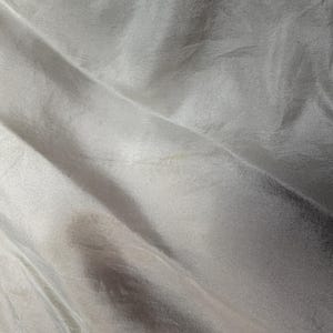 Op de afbeelding: Een stuk lichtgrijze stof met een glad, reflecterend oppervlak en een subtiele glans. Het materiaal vertoont zachte, vloeiende plooien en diepe vouwen die een dynamisch spel van licht en schaduw cre&euml;ren over het golvende oppervlak.