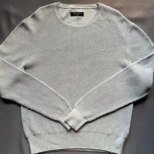 May include: A light gray waffle knit sweater with a small logo on the chest. The sweater has long sleeves and a crew neck.