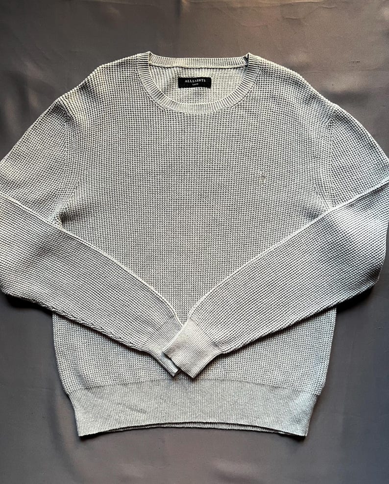 May include: A light gray waffle knit sweater with a small logo on the chest. The sweater has long sleeves and a crew neck.