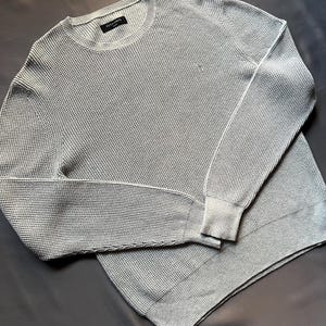 May include: A light gray waffle knit sweater with a crew neck and long sleeves. The sweater has a small label on the front that says "ALLSAINTS".