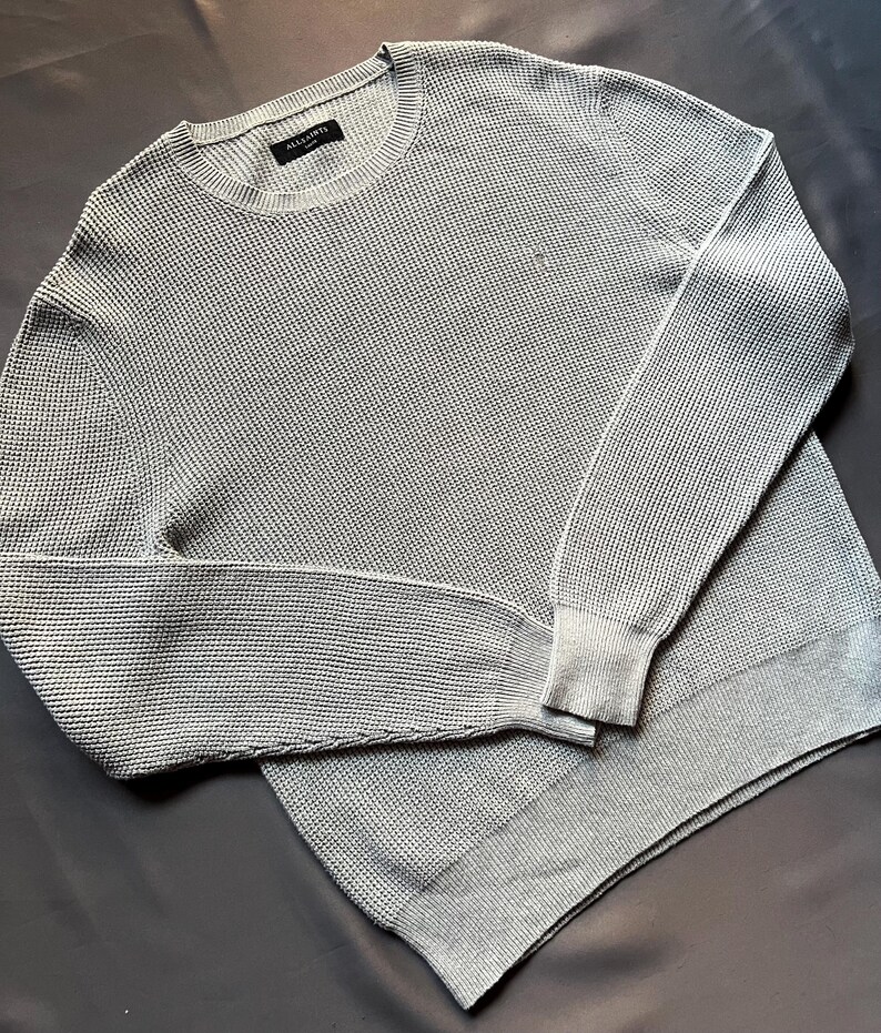 May include: A light gray waffle knit sweater with a crew neck and long sleeves. The sweater has a small label on the front that says "ALLSAINTS".