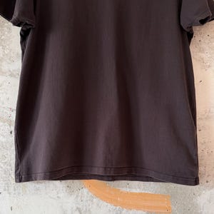 May include: A dark brown t-shirt with short sleeves. The shirt is made of a soft, slightly textured fabric. The background is a light-colored wall with a painted design.