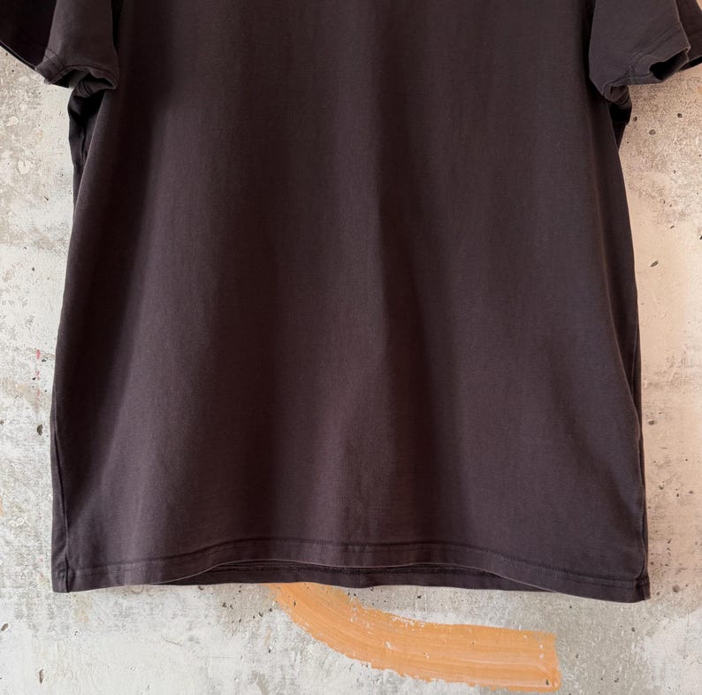 May include: A dark brown t-shirt with short sleeves. The shirt is made of a soft, slightly textured fabric. The background is a light-colored wall with a painted design.