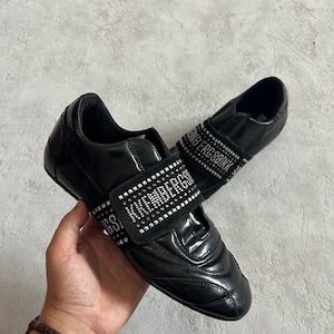 May include: A pair of black sneakers with a glossy finish, featuring a wide strap across the top adorned with silver studs and the text BIKKEMBERGS. The shoes have a rounded toe, a low-top silhouette, and a textured black fabric strap detail.