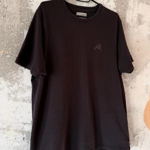May include: A black t-shirt with short sleeves hangs on a black hanger. The shirt has a crew neck and a small, embroidered logo on the chest. The background is a textured, off-white wall.