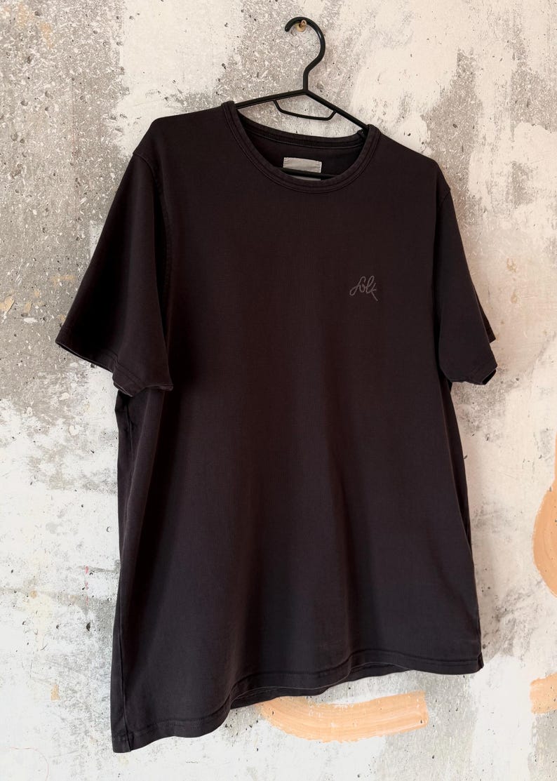 May include: A black t-shirt with short sleeves hangs on a black hanger. The shirt has a crew neck and a small, embroidered logo on the chest. The background is a textured, off-white wall.