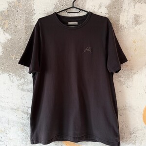 May include: A black crew neck t-shirt hanging on a black hanger. The shirt has short sleeves and a small embroidered design on the left chest. The background is a textured, off-white wall with gray and orange accents.