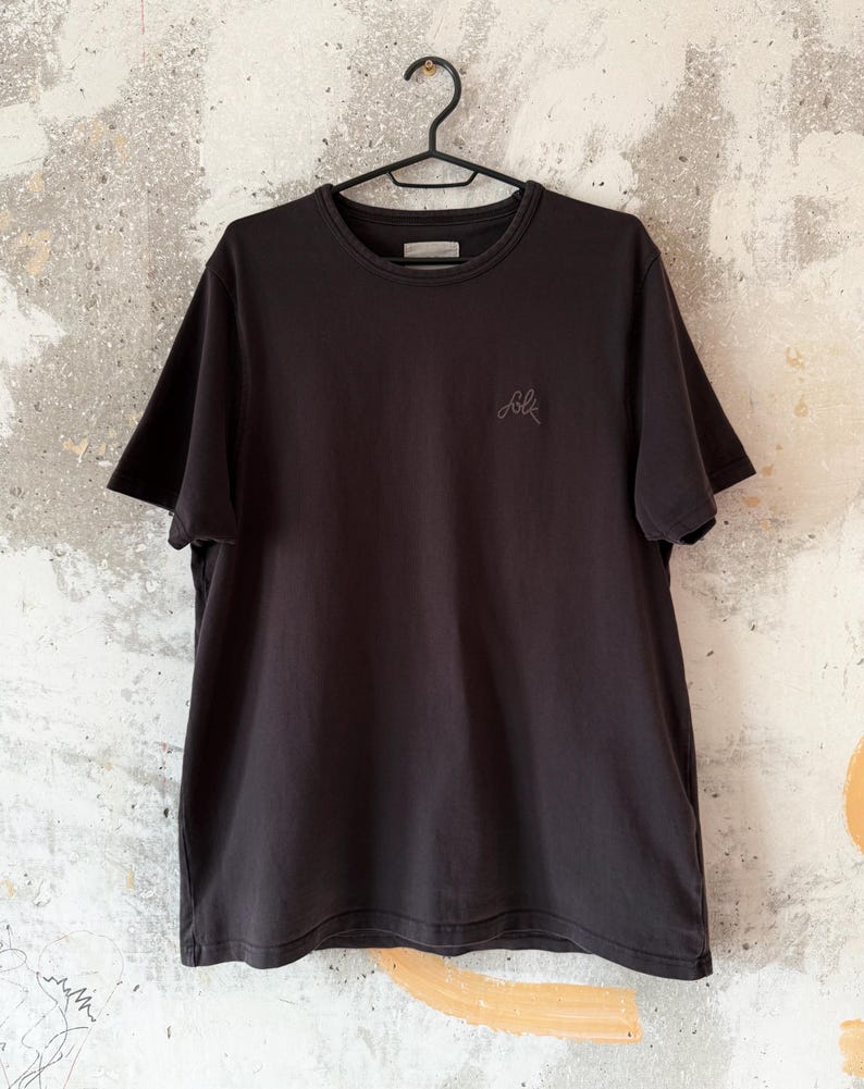 May include: A black crew neck t-shirt hanging on a black hanger. The shirt has short sleeves and a small embroidered design on the left chest. The background is a textured, off-white wall with gray and orange accents.