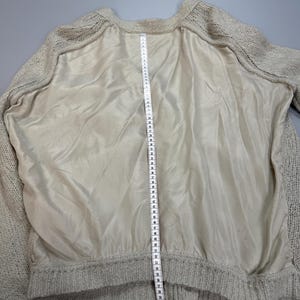 Op de afbeelding: Een beige gebreide trui met een glad, effen middenpaneel en mouwen van gebreide textuur. Een wit meetlint is verticaal over het midden geplaatst en toont een lengtemeting die op het lint voorbij de 64 cm reikt.