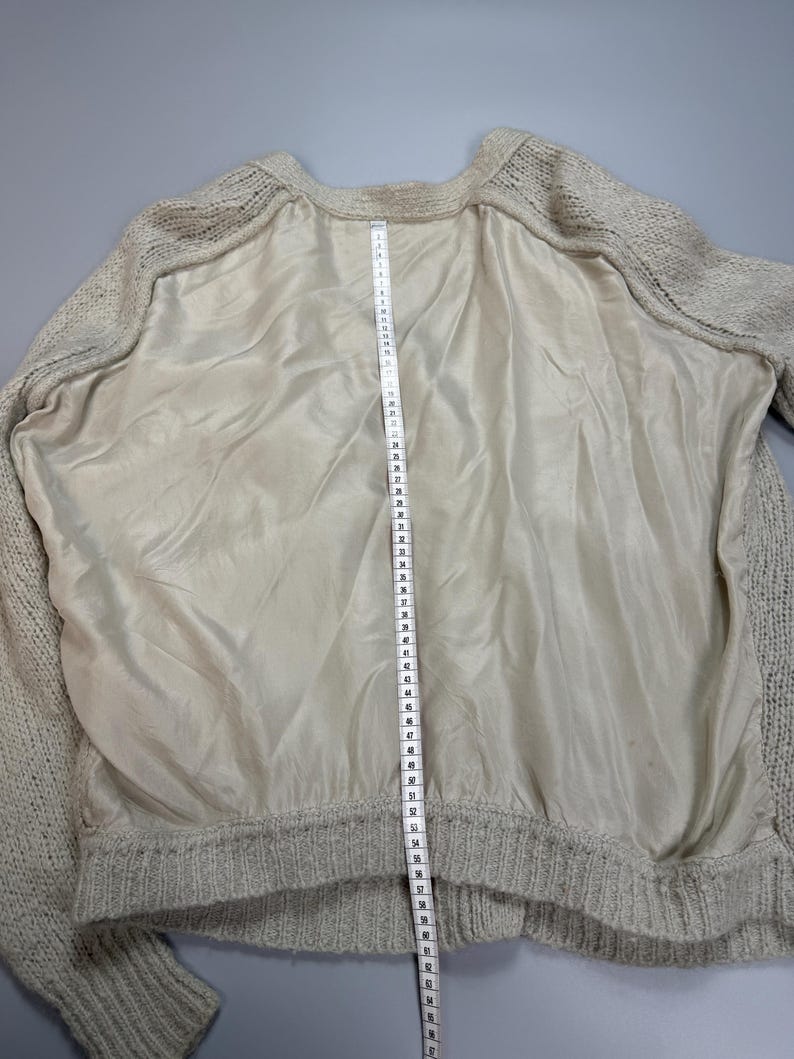 Op de afbeelding: Een beige gebreide trui met een glad, effen middenpaneel en mouwen van gebreide textuur. Een wit meetlint is verticaal over het midden geplaatst en toont een lengtemeting die op het lint voorbij de 64 cm reikt.