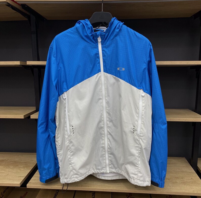 K&ouml;nnte beinhalten: Eine blaue und wei&szlig;e leichte Jacke mit Rei&szlig;verschluss und Kapuze. Die Jacke hat ein Logo auf der Brust, das "Oakley" sagt.