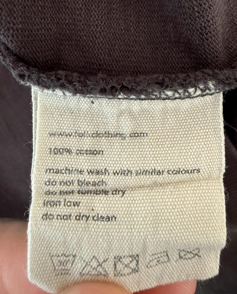 May include: Close-up of a clothing tag with the text "www.folkclothing.com" and "100% cotton." The tag also includes washing instructions: machine wash with similar colors, do not bleach, do not tumble dry, iron low, and do not dry clean.