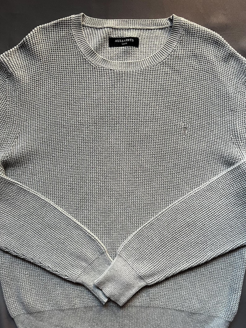 May include: A light gray waffle knit sweater with a crew neck and long sleeves. The sweater has a small "AllSaints" label on the front.