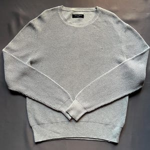 May include: A light gray waffle knit sweater with long sleeves and a crew neck. The sweater has a small logo on the front.