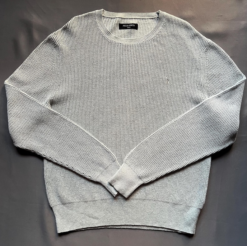 May include: A light gray waffle knit sweater with long sleeves and a crew neck. The sweater has a small logo on the front.