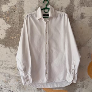 May include: A white long-sleeve button-down shirt hanging on a green hanger. The shirt has a classic collar and buttoned cuffs. The shirt is made of a smooth fabric.