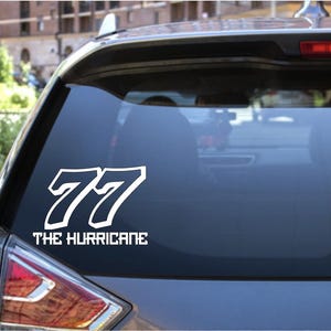 May include: White vinyl decal applied to a car's rear window. The decal features the number "77" in a bold, stylized font, with the words "THE HURRICANE" below. The car is a light blue color.