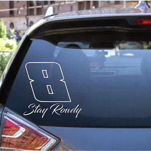 Könnte beinhalten: Weißer Vinyl-Aufkleber auf einer Autoscheibe. Der Aufkleber zeigt eine stilisierte Zahl 8 mit dem Schriftzug "Stay Roudy" in Schreibschrift. Das Rücklicht des Autos ist sichtbar.