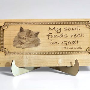 May include: Wooden plaque featuring a sleeping kitten illustration and the text "My soul finds rest in God! Psalm 62:1". The plaque is supported by a wooden stand. The design is engraved on the light-coloured wood.
