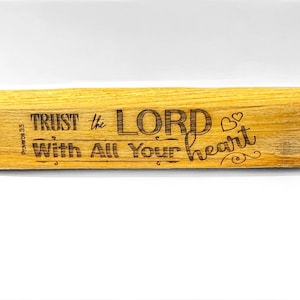 May include: A rectangular wooden block with the engraved text "TRUST the LORD With All Your heart". The wood has a light brown colour and the text is in a dark brown colour. The block is a decorative item.