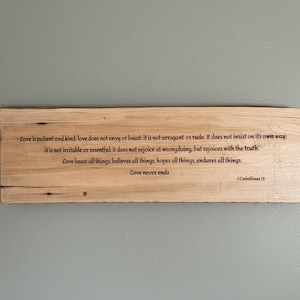 Puede incluir: Un letrero rectangular de madera clara con texto grabado de 1 Corintios 13. El letrero está montado en una pared gris. El texto está en una fuente sencilla y elegante.