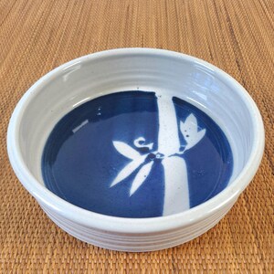 May include: A round ceramic bowl with a light gray exterior and a deep blue interior. The bottom of the bowl features a white, stylized graphic of a bamboo stalk with leaves. The bowl has a smooth, glossy finish and a slightly ribbed outer wall.