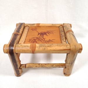 May include: A small, square bamboo stool with a natural finish. The top features a bamboo leaf design. The legs and frame are constructed from bamboo poles, with darker brown accents on the joints and edges. A decorative piece for home decor.