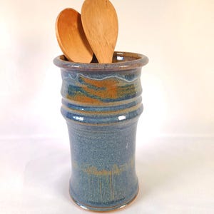 May include: A handmade blue and brown ceramic utensil holder with two wooden spoons. The cylindrical holder has a textured surface and a flared rim. The spoons are light brown and partially visible.
