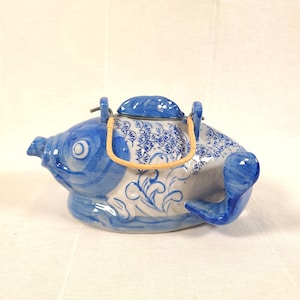 May include: A ceramic teapot shaped like a fish, painted in blue and white. The teapot has a lid, a spout, and a handle made of woven material. The fish design includes scales and decorative swirls.