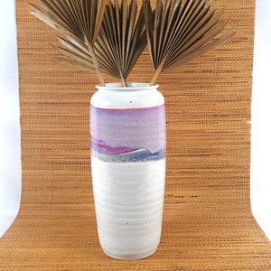 May include: A tall, white ceramic vase with a textured, ridged base and a painted upper section. The upper section features a gradient of colors, including purple, pink, and blue. Dried palm leaves are placed inside the vase.