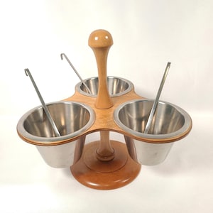 May include: A wooden condiment server with three stainless steel bowls and small spoons. The server has a light wood base and a central wooden handle. The bowls are arranged in a triangular pattern, perfect for serving dips or sauces.