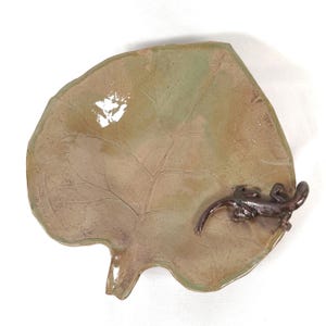 May include: A ceramic leaf-shaped dish with a brown and green glaze. The dish features a detailed leaf vein pattern and a small, dark brown lizard figurine on the edge. The dish is designed for decorative use.