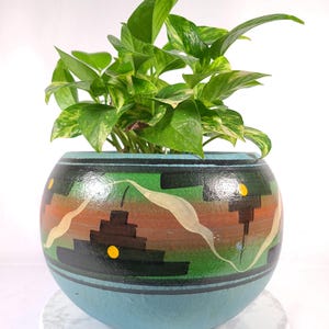 May include: A round, light blue planter featuring a hand-painted design of black stepped geometric shapes, horizontal brown and green bands, and small yellow dots. A green and yellow variegated pothos plant grows from the top of the pot.