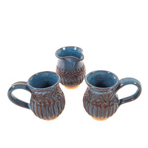 May include: A set of three ceramic pieces including two mugs with side handles and one small pitcher, all featuring a deep blue glaze with a dark brown textured pattern of vertical lines and geometric shapes, set against a plain white background.