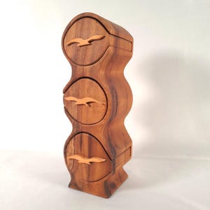 May include: A wooden jewelry box with three drawers, each adorned with a carved bird design. The box has a curved, organic shape and is made of a warm, reddish-brown wood. The birds are a lighter shade of brown.