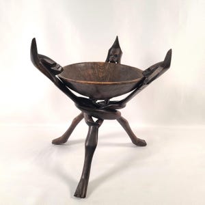 May include: A dark brown, hand-carved wooden bowl on a tripod base. The bowl is supported by three figures with outstretched arms. The legs of the stand are carved to resemble human legs. The bowl is approximately 10 inches in diameter.