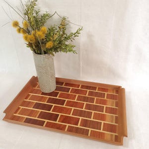 May include: A rectangular wooden serving tray with a brick pattern design. The tray is made of various shades of brown wood, with lighter wood accents. A vase with yellow and green flowers sits on the tray.