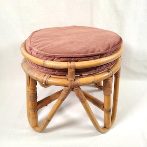 May include: A round, vintage rattan stool with a plush, dusty rose cushion. The stool features a woven rattan frame with curved legs and a circular seat. The cushion is made of a textured fabric. The stool is approximately 16 inches tall.