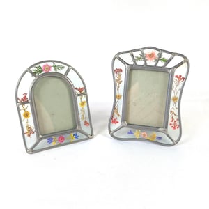 May include: Two standing picture frames constructed with silver-toned metal borders and clear glass panels. Each frame features pressed, dried flowers in shades of pink, yellow, red, and blue embedded within the glass borders around the central opening.