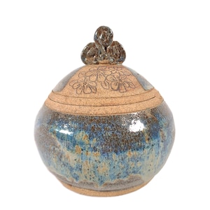 May include: A round ceramic jar with a lid featuring a sculpted handle. The body has a mottled blue and brown glaze with a glossy finish. The lid is unglazed, textured, and decorated with an etched floral pattern of five-petaled blossoms.